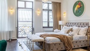 Une chambre à coucher avec une tête de lit capitonnée, une table basse en verre, un fauteuil vert et une vue sur l’extérieur grâce à une grande fenêtre.