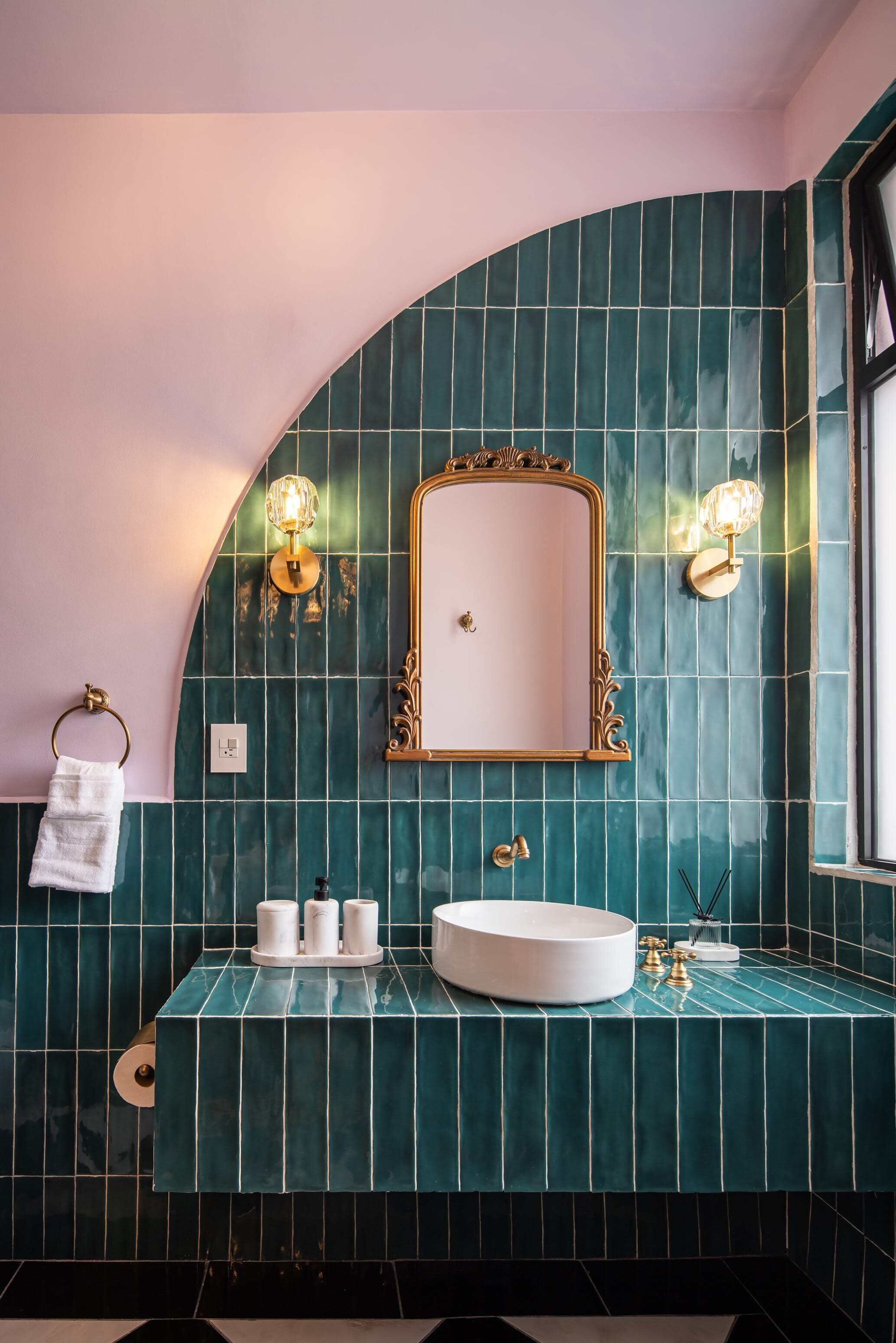 Un bany amb una paret corba, un lavabo rodó, un mirall amb una estructura ornamentada i dues làmpades subjectes a la paret.