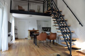 A modern loft apartment with a wooden floor, a dining area with two chairs, a kitchen with white cabinets, and a black spiral staircase.