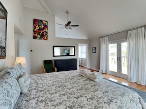A bedroom with a large bed, a ceiling fan, a dresser, a chair, a mirror, and a view of the outside through a sliding glass door.