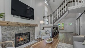 Un vaste salon doté d’une cheminée en pierre, d’un grand téléviseur à écran plat, d’une table basse en bois et d’un escalier menant à l’étage supérieur.