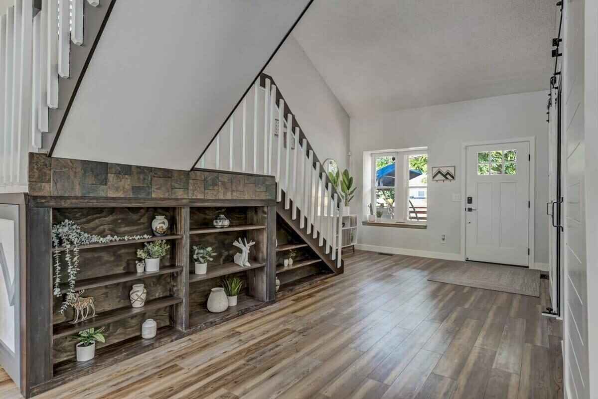 A spacious living area with a wooden staircase, a built-in shelf with decorative items, and a view of the outside.