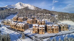 A resort with multiple chalet-style buildings, arranged in a semi-circular layout, surrounded by snow-covered trees and a mountain in the background. - 3BR Ski-In/Out Condo, Reno 2024, Explorer Gondola Access – Big Sky, MT (Big Sky)