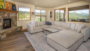 A spacious living room with a stone fireplace, large sectional sofa, and a view of a mountainous landscape through large windows. - 3BR Ski-In/Out Condo, Reno 2024, Explorer Gondola Access – Big Sky, MT (Big Sky)