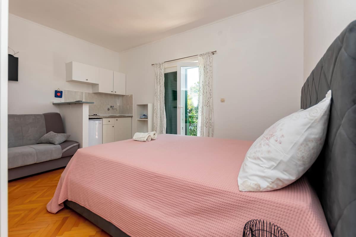 A bedroom with a bed, a grey sofa, a kitchen area, and a window with curtains.