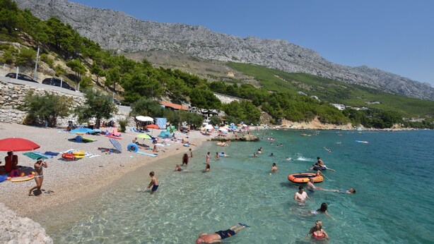A beach with clear water, people swimming and sunbathing, and a rocky hillside in the background.