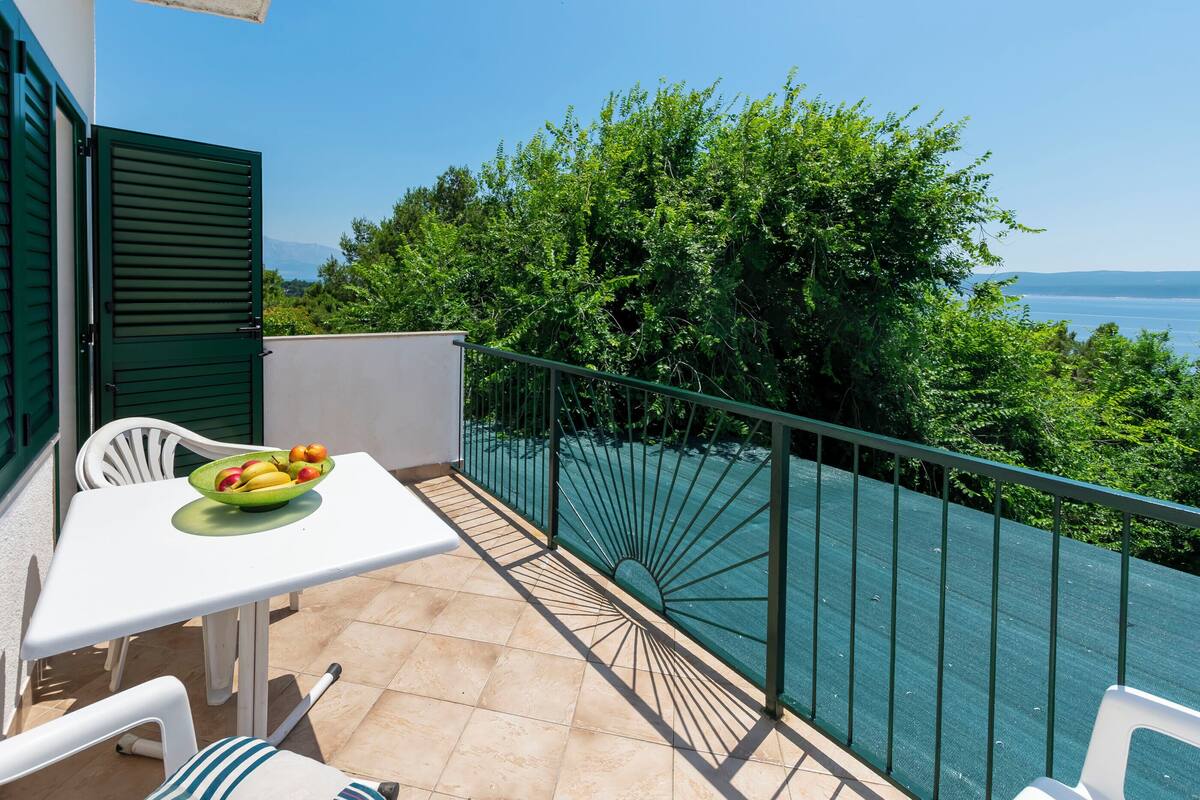 A balcony with a table and chairs, a bowl of fruit, and a view of the sea and greenery.