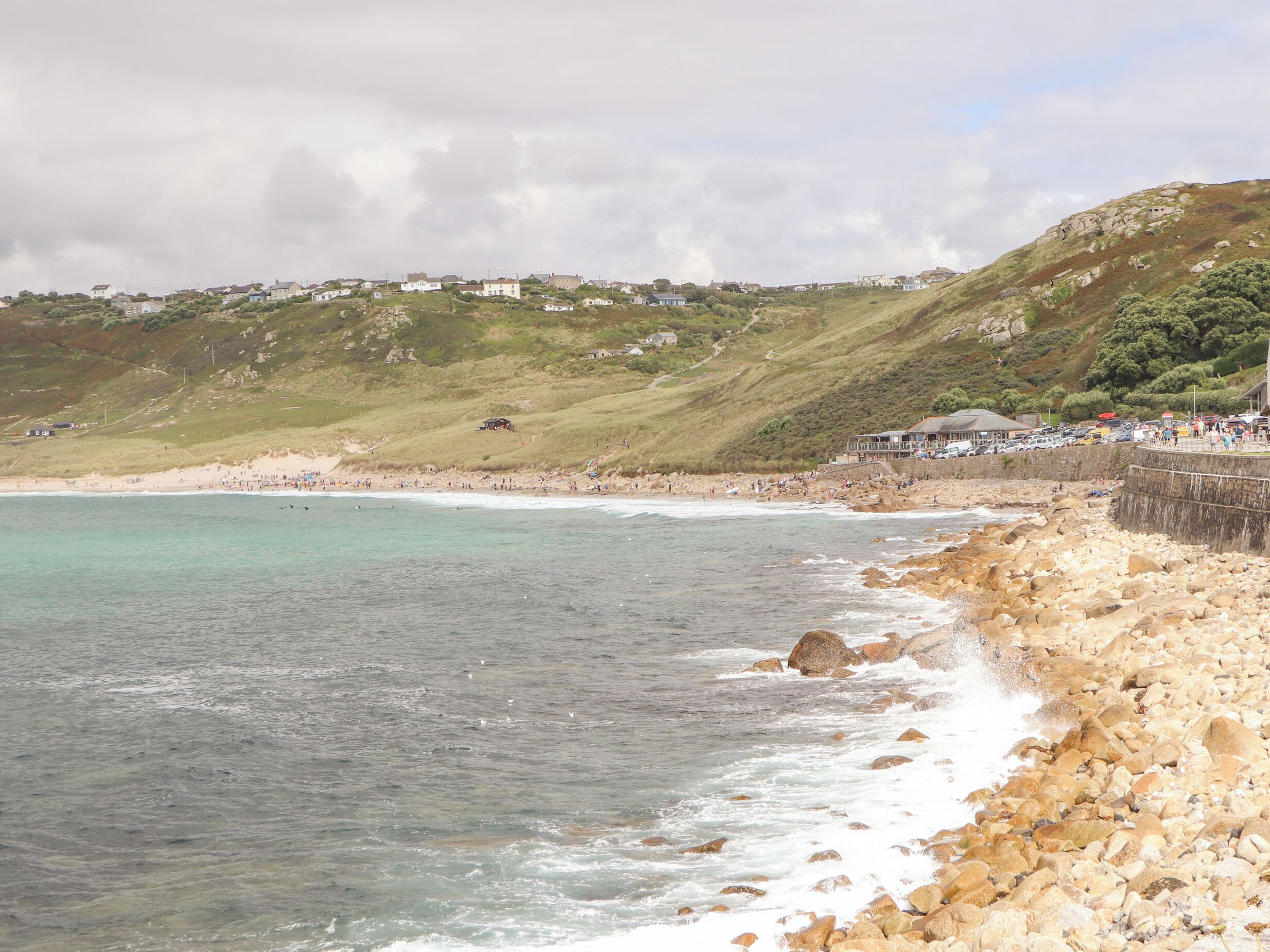 A coastal landscape with a rocky beach, a clear view of the sea, and a hillside with buildings.