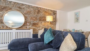 Una sala de estar con pared de piedra, un espejo redondo, un sofá con cojines, una lámpara y un comedor con mesa y sillas.