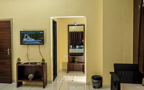 A hotel room with a TV mounted on the wall, a bed with a mirror above it, a small table with a plant, and a wicker chair. - THE SHRINE HOTEL (Khandwa)