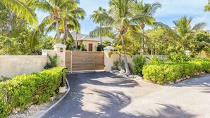 A gated residential area with palm trees, a paved driveway, and a house with a wooden gate. - Garden Escape Near Beach, Bikes and BBQ Included (Providenciales)