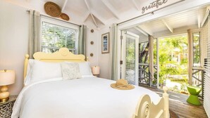 A bedroom with a bed, bedside lamps, a hat on the bed, and a balcony with a chair. - Garden Escape Near Beach, Bikes and BBQ Included (Providenciales)