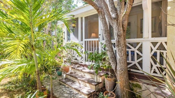 A covered porch with steps leading to a white railing, surrounded by potted plants and a large tree. - Garden Escape Near Beach, Bikes and BBQ Included (Providenciales)