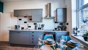A modern kitchen with grey cabinets, a stainless steel range hood, and a dining area set for six with a table, chairs, and glassware. - Apartment 3 - The Crescent (York)