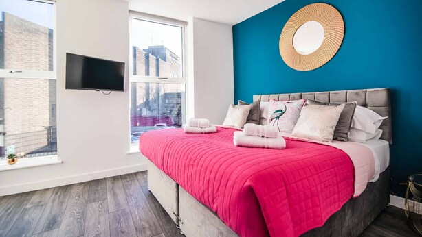 A modern bedroom with a large bed, a TV mounted on the wall, and a view of the city through large windows. - Apartment 3 - The Crescent (York)