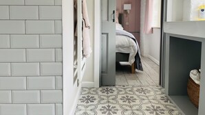 A bathroom with white tiled walls, a patterned floor, a bed with a white and gray bedspread, and a wicker basket on the right. - The Hayloft (Crewe)