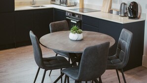 A modern kitchen with dark cabinetry, a round dining table with chairs, and a window with a blind. - Apartment 7 (York)
