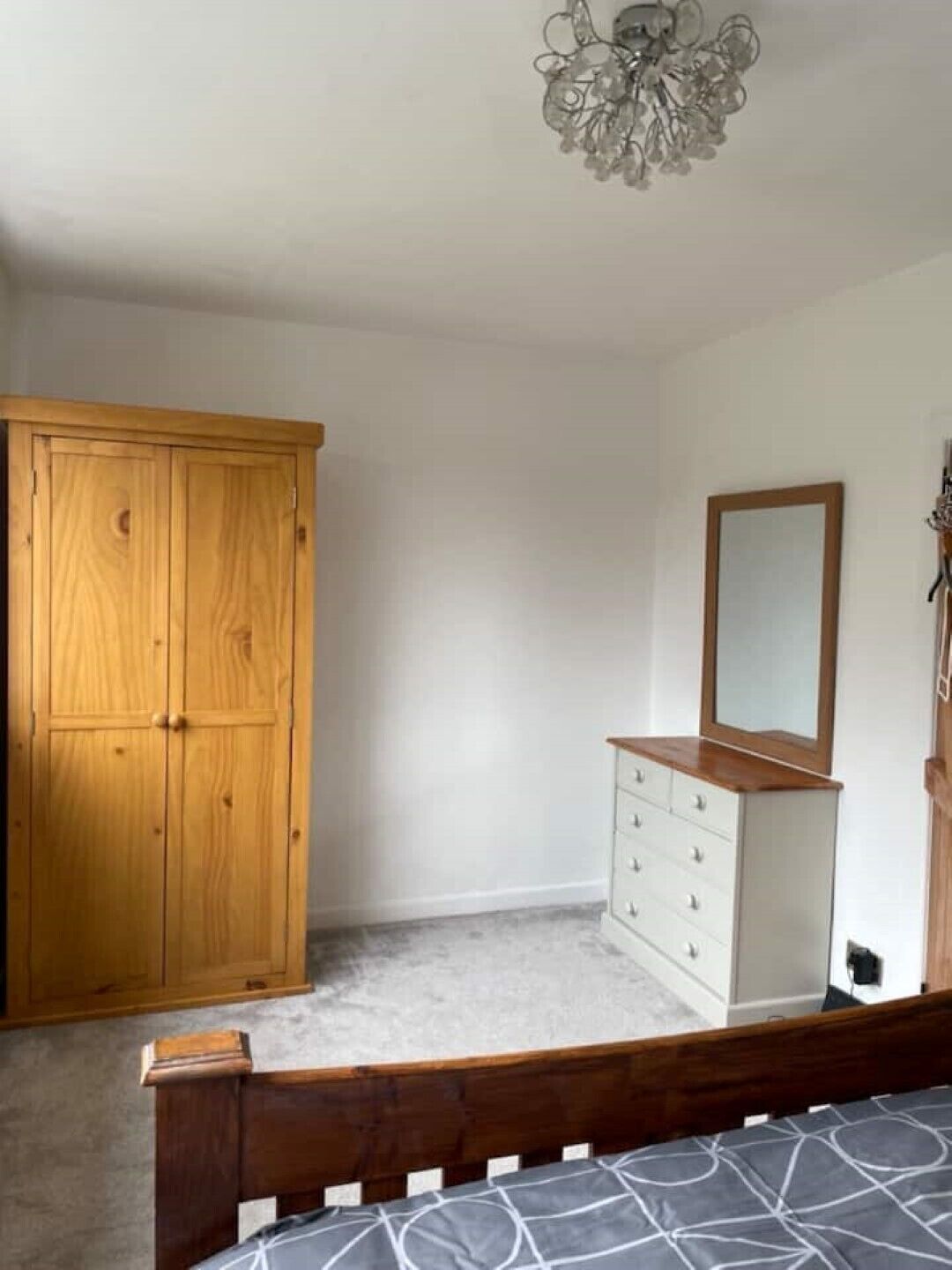 A bedroom with a wooden wardrobe, a dresser with drawers, a bed with a wooden headboard, and a chandelier.
