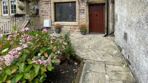 A stone building with a traditional design, featuring a front door, windows, and a small garden area with pink flowers.