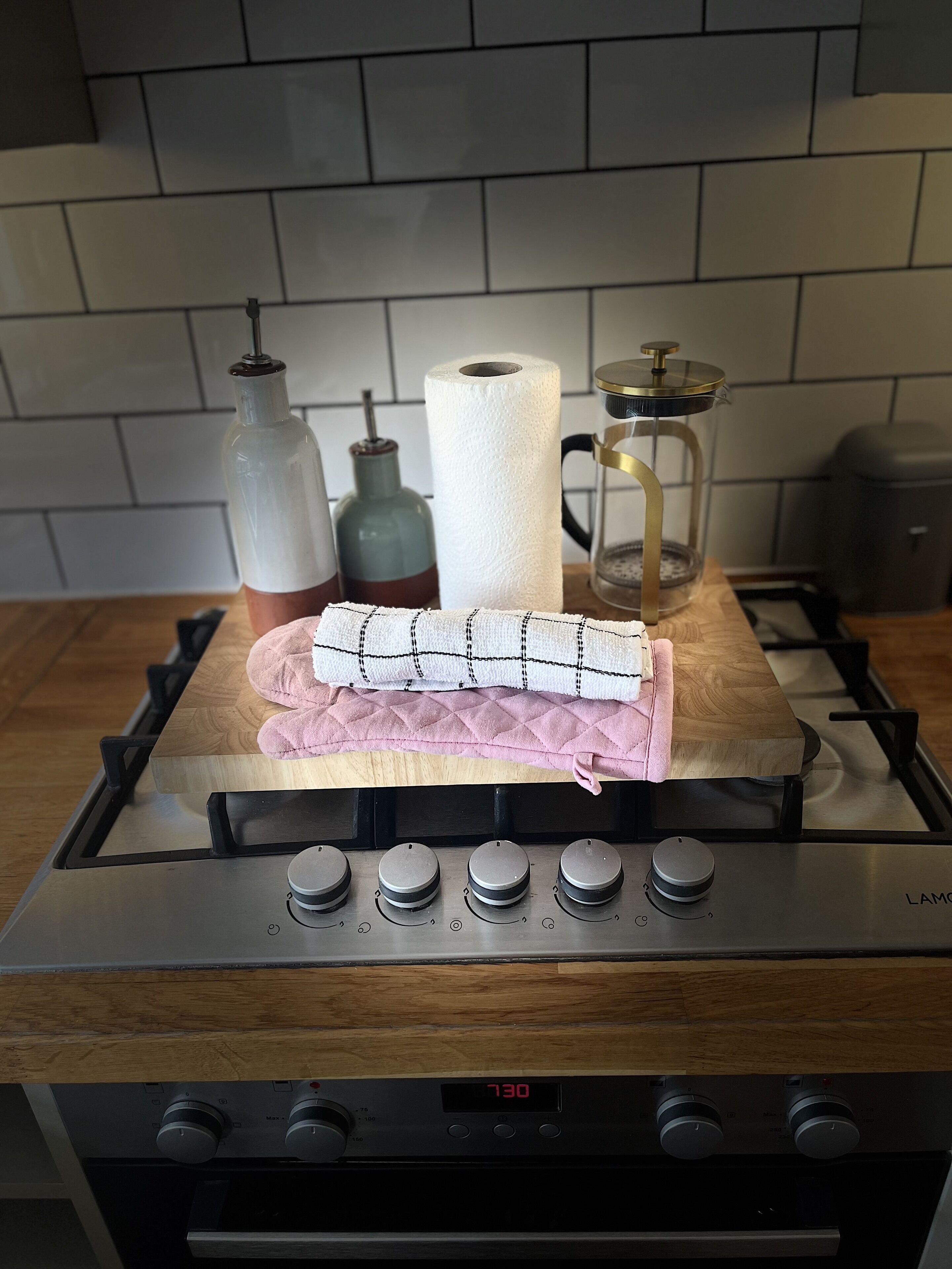 A kitchen with a gas stove, a wooden cutting board, a pink towel, a roll of paper towels, a glass jar, and a metal container.