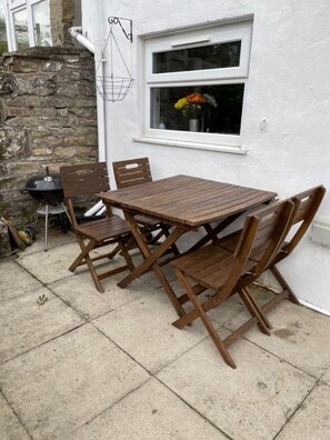 A patio with a wooden table and chairs, a barbecue grill, and a window with flowers. - Spring Cottage 2BR Escape - Garden, Coffee Machine, Self Check-in (Trawden)