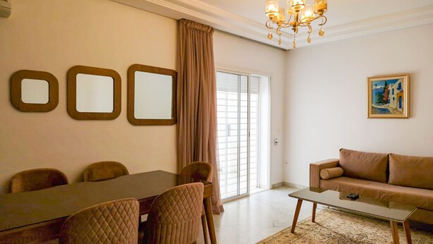 A living room with a dining table, chairs, a sofa, a coffee table, a chandelier, and a painting on the wall. - Ennaser Bliss Your Urban Oasis Awaits (Ariana)