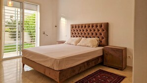 A bedroom with a large bed, a nightstand, a lamp, and a view of greenery through the window. - Ennaser Bliss Your Urban Oasis Awaits (Ariana)