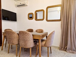 A dining area with a wooden table and four cushioned chairs, a flat-screen TV mounted on the wall, and three framed mirrors. - Ennaser Bliss Your Urban Oasis Awaits (Ariana)