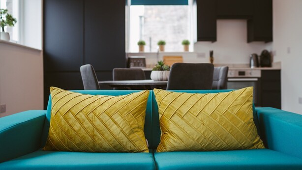A teal sofa with two yellow pillows in a modern living room with a kitchen in the background. - Apartment 8 (York)
