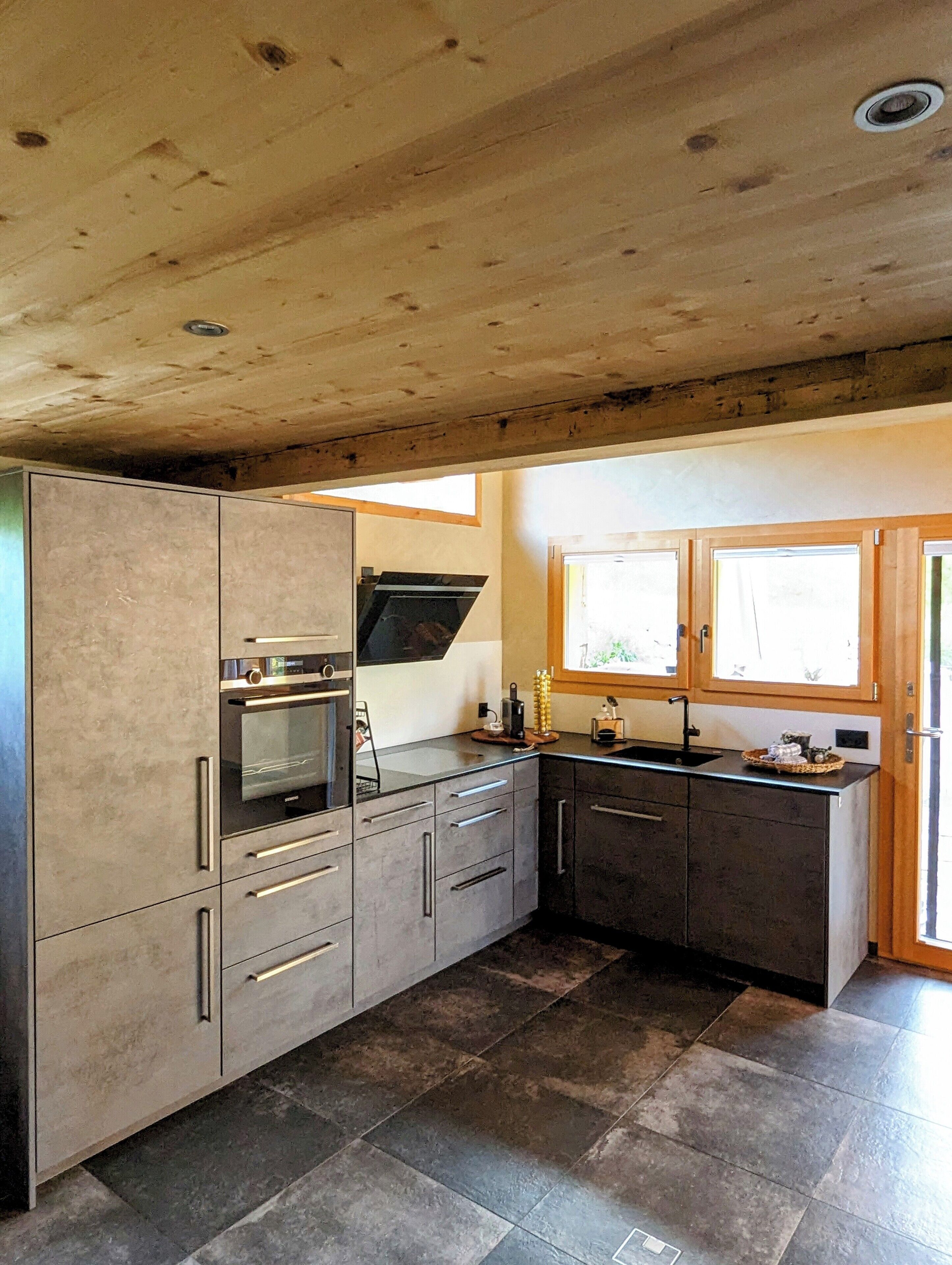 Une cuisine moderne équipée d’un four encastré, d’un four à micro-ondes et d’un évier. Une fenêtre se trouve au-dessus de l’évier et le plafond est en bois.
