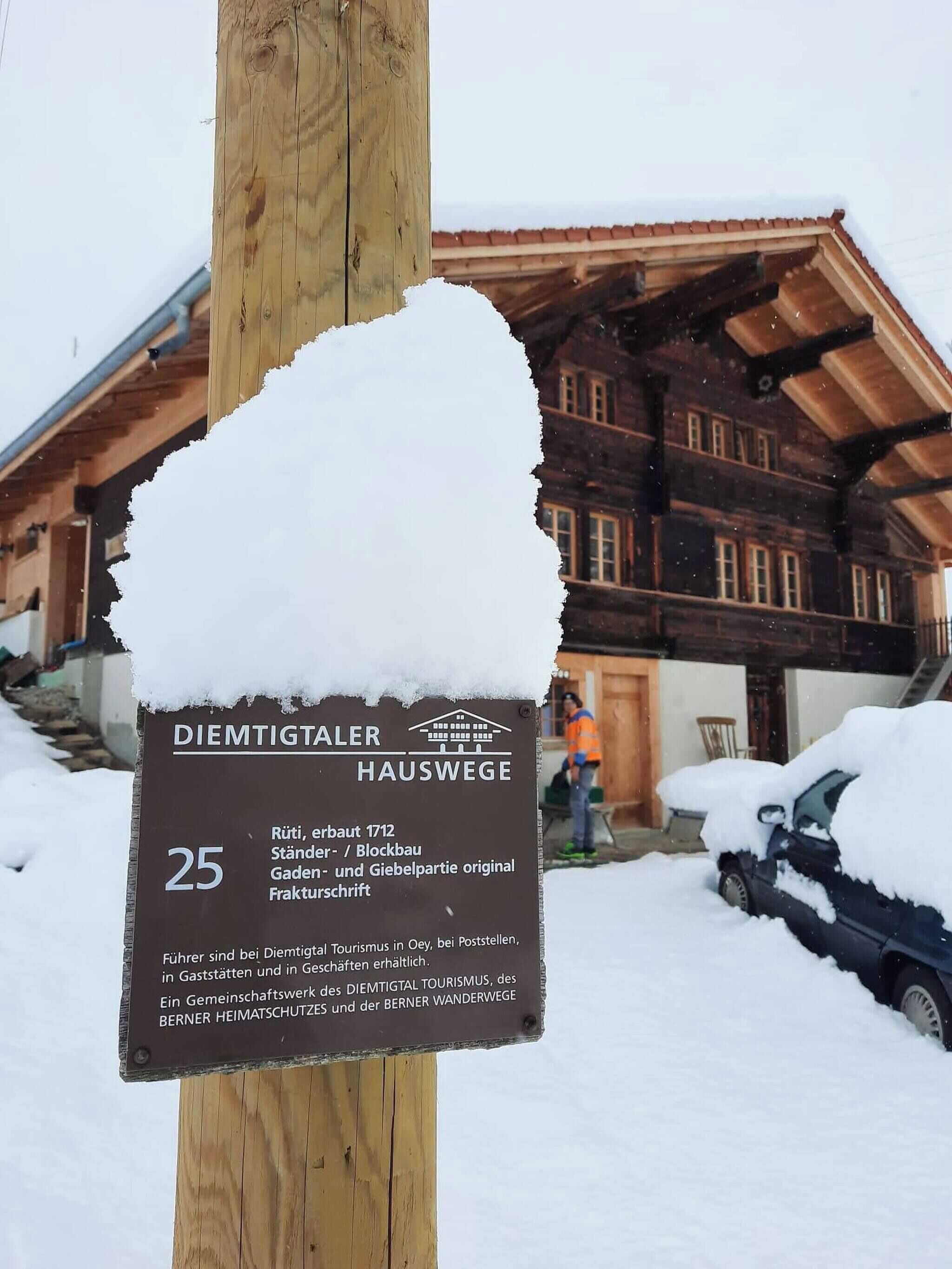 Segnaletica per DIEMTIGTALER HAUSWEGE, con un palo di legno, paesaggio innevato e un edificio tradizionale in legno sullo sfondo.