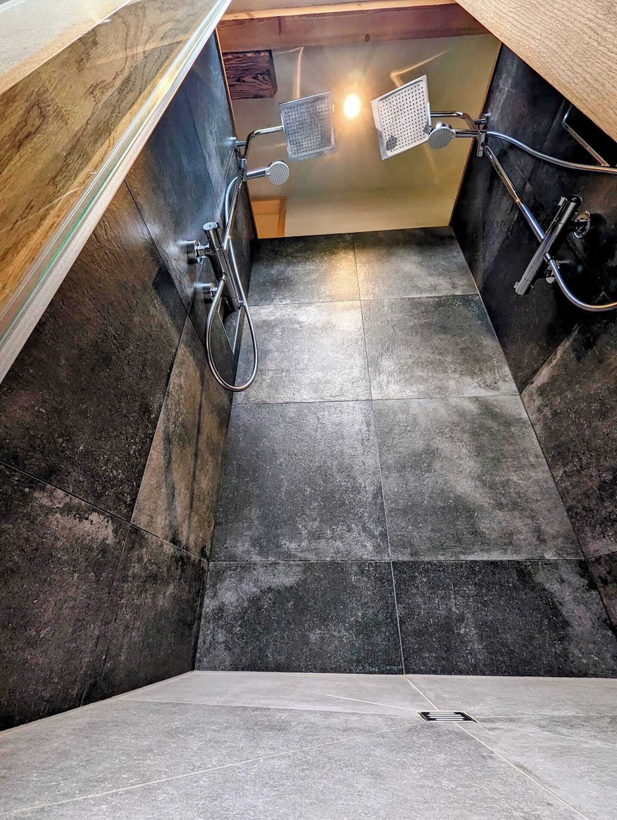 Un estrecho pasillo con baldosas y una zona de ducha con ducha de cabeza metálica y rociador de mano.