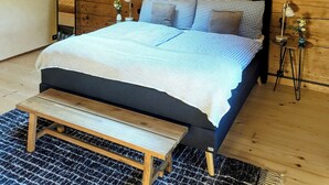 Quarto com claraboia, cama com roupa de cama xadrez, mesa de cabeceira com abajur e um banco.