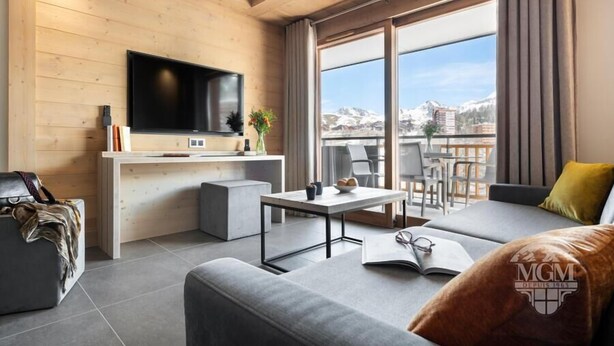 A modern living room with a flat-screen TV, a sofa, a coffee table, and a balcony with a view of mountains. - Wifi, ski-in/ski-out & foot of the slopes, view resort, fitness, swimming pool, sauna, hammam (La Plagne)