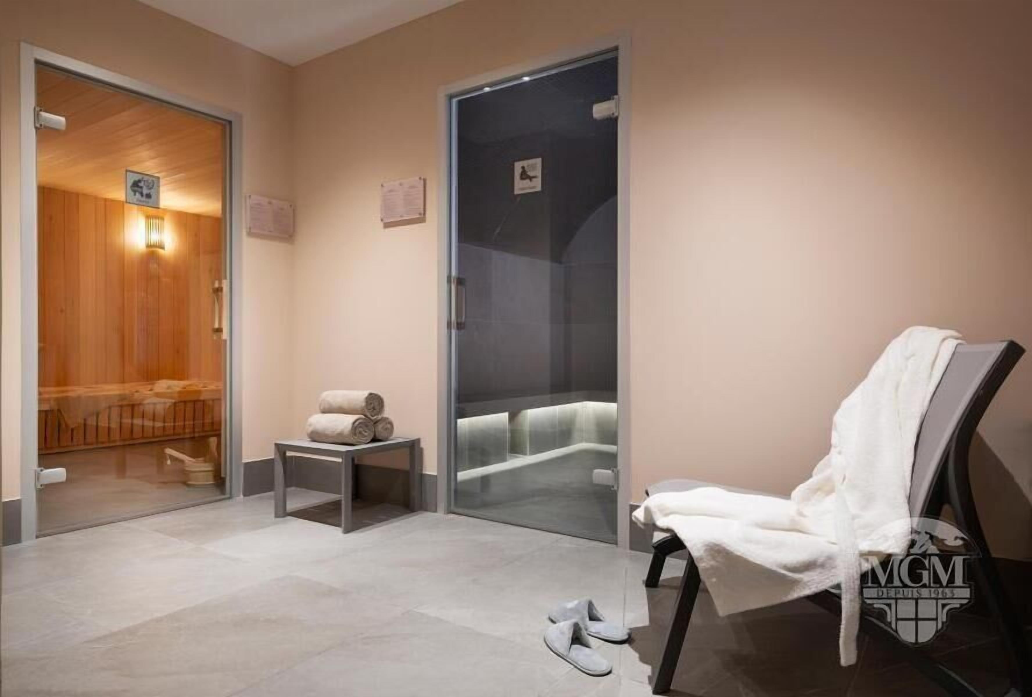 A spa room with a wooden door, a glass partition, a massage table, a towel, and slippers on the floor.