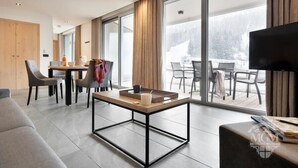 Una habitación de hotel moderna con un gran ventanal corredera que da a un balcón, un comedor y una zona de estar con una televisión de pantalla plana.