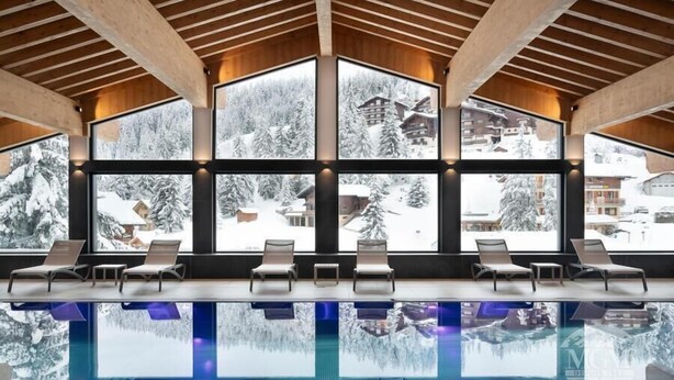 Une vaste piscine intérieure à l’eau claire, avec des poutres en bois au plafond et de grandes fenêtres offrant une vue imprenable sur les montagnes enneigées.
