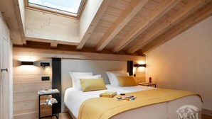 Une chambre avec un puits de lumière, des poutres en bois, un lit avec une literie blanche et jaune, des tables de chevet et une petite table de nuit.