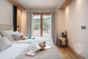 Camera da letto con un letto grande, un comodino con un orsacchiotto, un libro e vista su un balcone con tende.