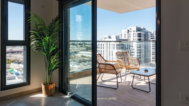 Un balcón con sillas de mimbre, una mesa, una planta en maceta y vistas a edificios.