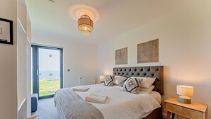 A bedroom with a large bed, a bedside table with a lamp, a chair, and a view through an open door. - Cottage 18 - 3 Bed (Pwllheli)