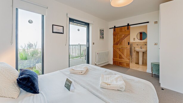 A bedroom with a large bed, a view to the outside, a wooden door, and a bathroom with a sink and mirror. - 2 Bed Ground Floor Apartment - 11 (Pwllheli)