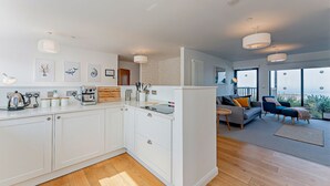 A modern kitchen with white cabinets, a coffee machine, and a wooden cutting board. There is a dining area with a sofa and chairs, and a large window offering a view of the outdoors. - 2 Bed Ground Floor Apartment - 11 (Pwllheli)