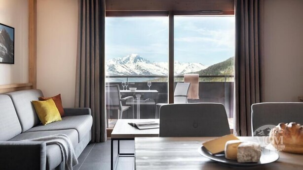A modern room with a grey sofa, a dining table with a cheese platter, and a view of snow-capped mountains through large windows. - Wifi, ski-in/ski-out & foot of the slopes, view mountain, fitness, swimming pool, sauna, hammam (La Plagne)