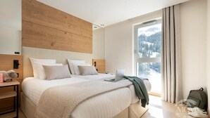 Un dormitorio moderno con una cama grande, una laptop sobre la cama, un cabecero de madera, una mesita de noche con una lámpara y vistas a un paisaje nevado a través de una ventana.