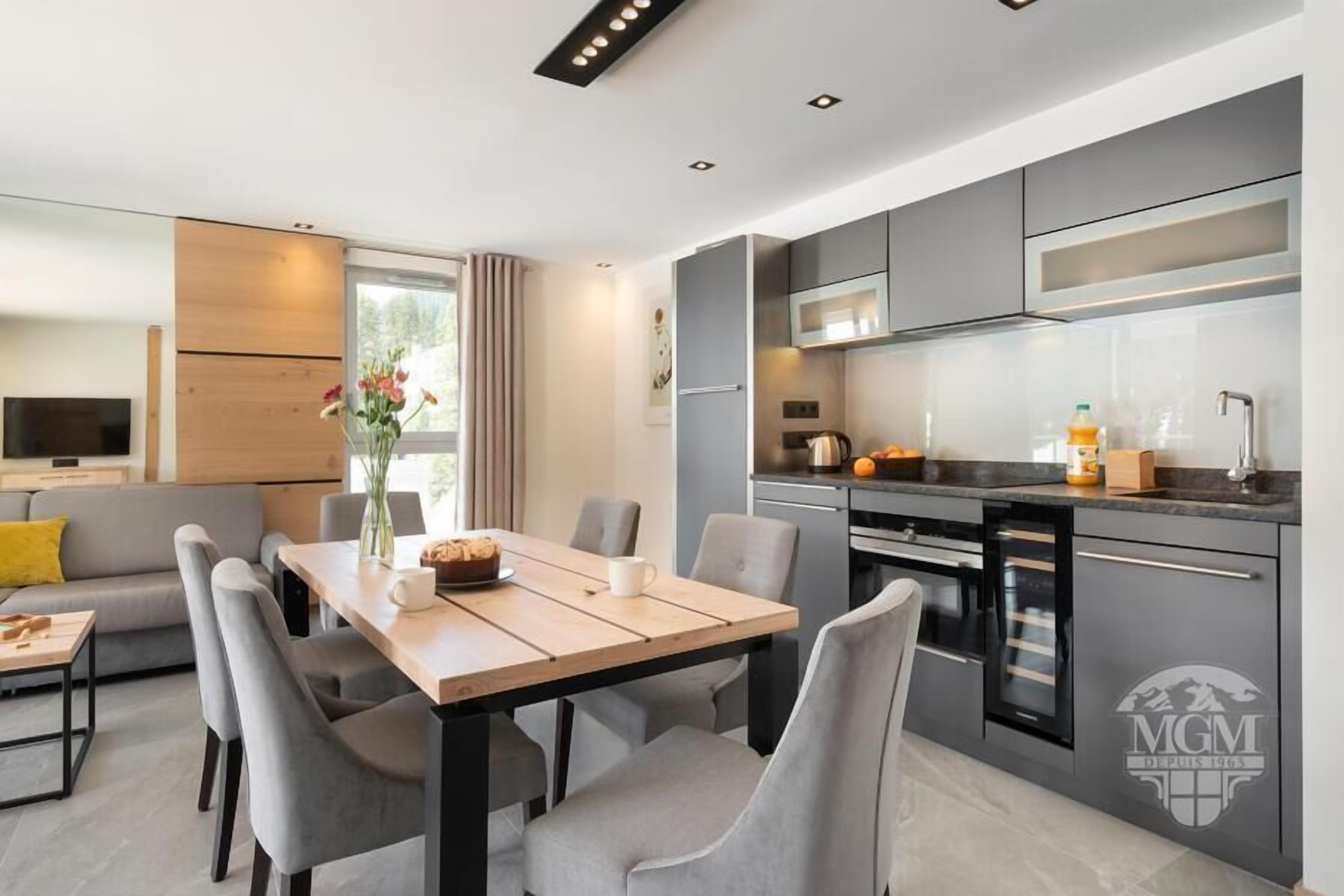 A modern kitchen with grey cabinets, a microwave, and a sink. There is a dining area with a table and chairs, and a sofa in the background.