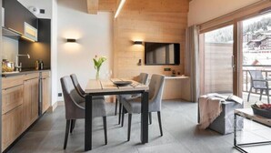 A modern kitchen with wooden cabinets, a dining table with chairs, a TV, and a view of a snowy landscape through large windows. - Wifi, ski-in/ski-out & foot of the slopes, fitness, swimming pool, sauna, hammam, balcony, tv, 45m² (Manigod)