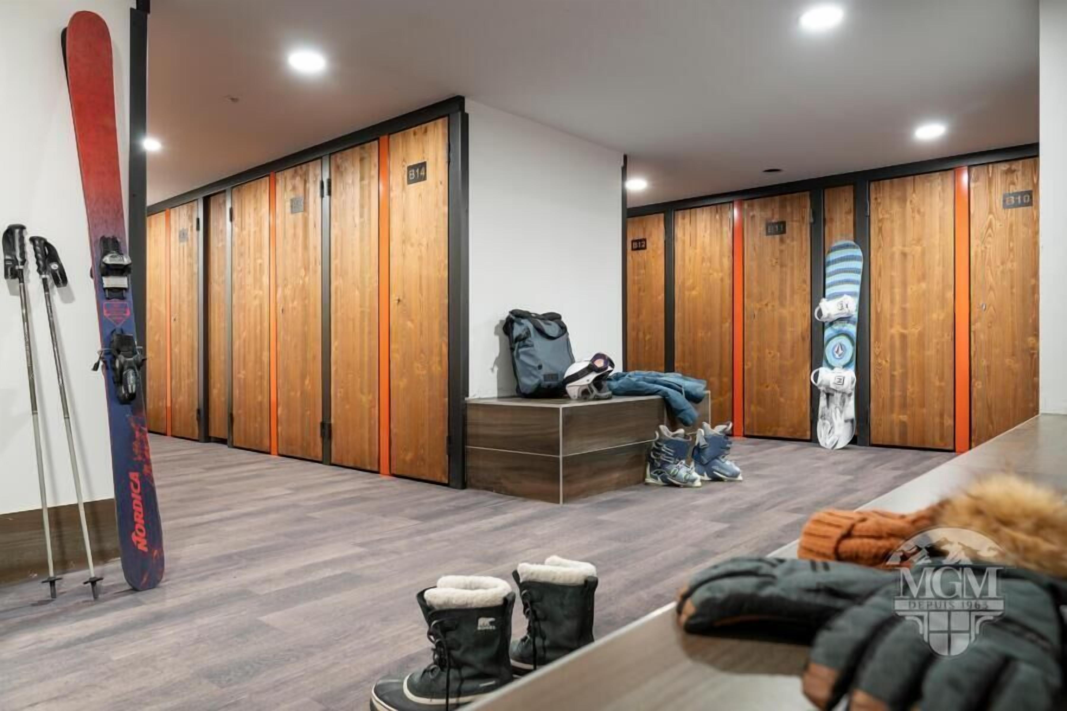 A hotel room with wooden lockers, a ski board leaning against the wall, and a bench with a backpack and shoes.