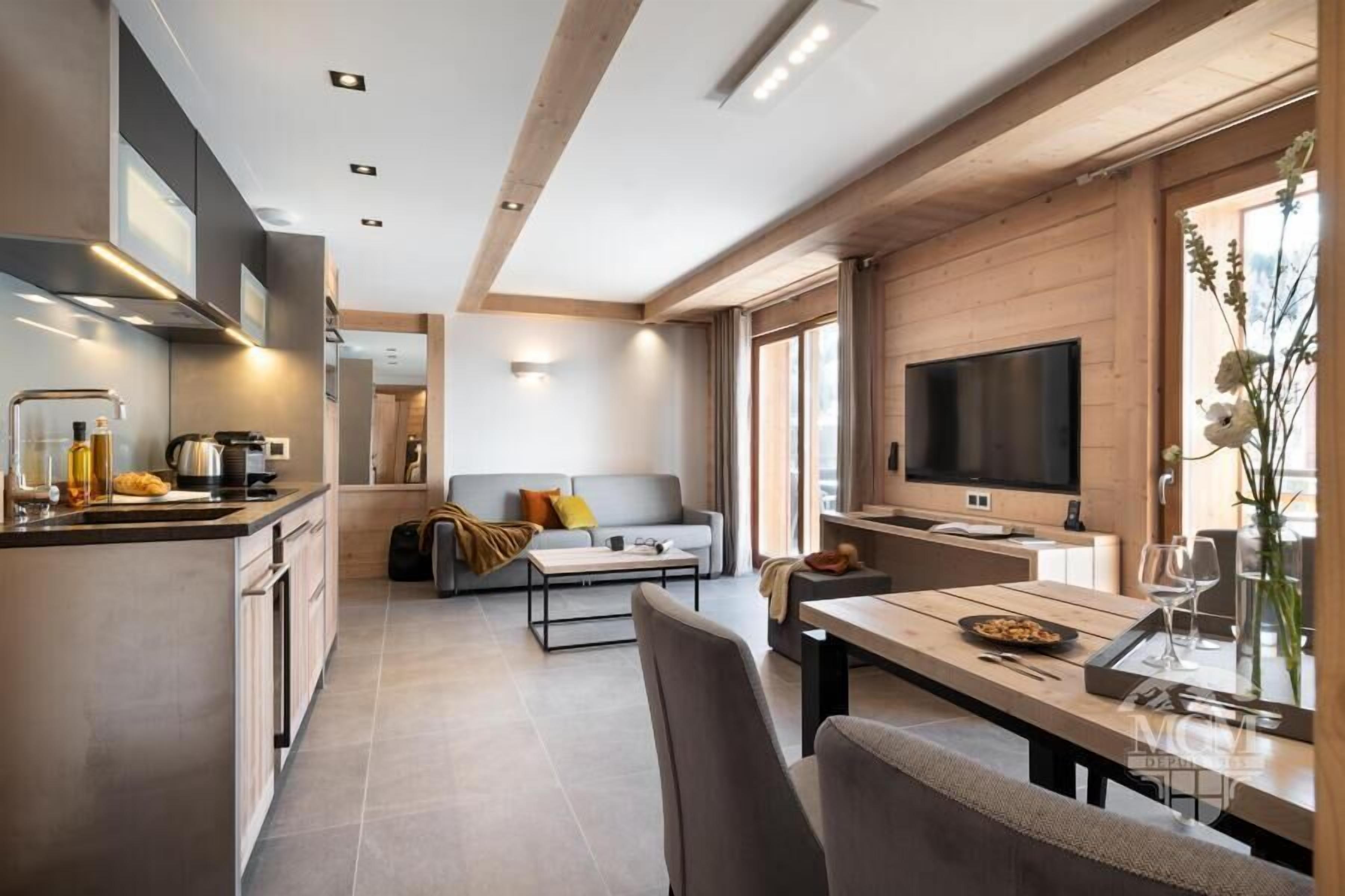 A modern kitchen with a central island, a dining area with a wooden table, and a living space with a sofa and TV.