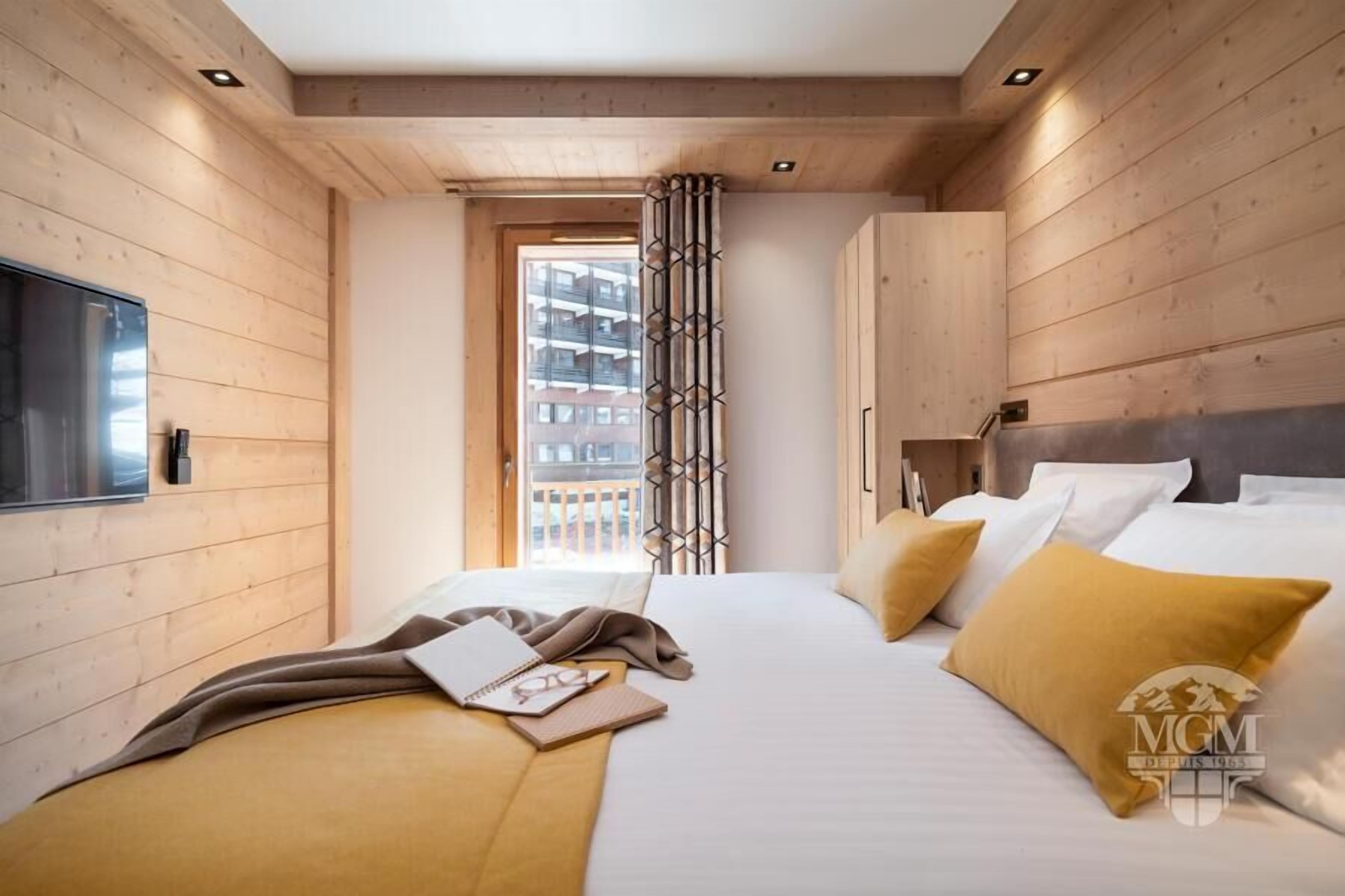A hotel room with a wooden interior, a bed with white and mustard pillows, a TV on the left wall, and a window with a view of buildings.